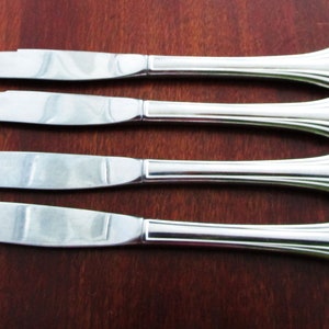 May include: Four silver butter knives with a decorative handle design. The blades are smooth and curved, and the handles have a fluted, fan-like shape. The knives are arranged on a dark wooden surface.