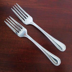 May include: Two stainless steel forks with four tines each. The handles have a teardrop shape with a beaded edge. The forks are placed diagonally on a dark brown wooden surface.