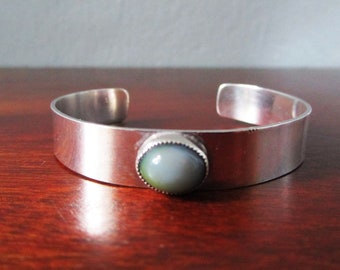 May include: A silver bangle bracelet with a round, iridescent green and gray stone set in the center.