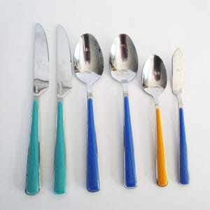 May include: A set of six stainless steel utensils: two knives with teal handles, two spoons with blue handles, a smaller spoon with an orange handle, and a butter knife with a blue handle. The utensils are arranged on a white surface.