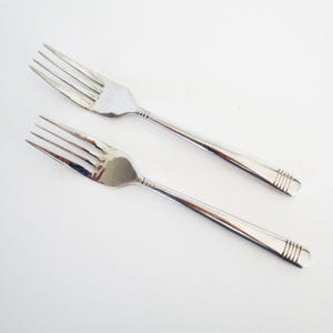 May include: Two matching stainless steel dinner forks with a polished silver finish. Each fork features four tines and a slender handle decorated with a series of three horizontal engraved lines near the rounded end of the handle base.