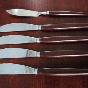 May include: Set of five stainless steel knives with brown wooden handles. The knives have serrated blades and are arranged in a row on a wooden surface.