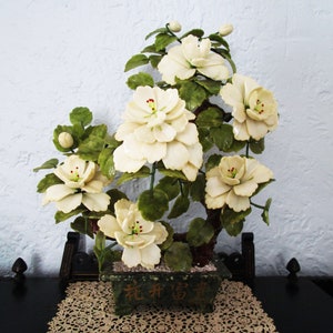May include: A white jade flower arrangement in a green jade pot with the Chinese characters "花开富贵" (hua kai fu gui) meaning "flowers bloom, wealth and prosperity" on the front. The arrangement is on a lace tablecloth.