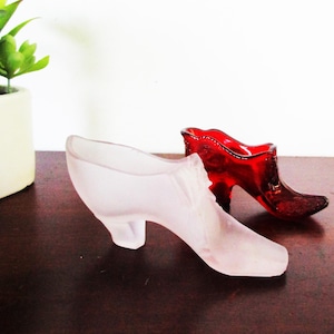 Puede incluir: Dos figuras de zapatos de tacón de cristal. Uno es rosa esmerilado, el otro es rojo vibrante. Ambos tienen un diseño de tacón clásico con punta y tacón curvo. Se exhiben sobre una superficie marrón oscuro.