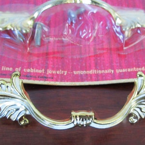 Può includere: Maniglia per armadio color oro con un design a foglie ornate. La maniglia è nella sua confezione originale, che include il testo "Cardware the prestige line of cabinet jewelry -- unconditionally guaranteed."