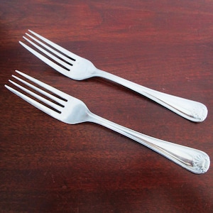 May include: Two stainless steel forks with four tines each, resting on a dark wood surface. The handles have a decorative design at the end. The forks are angled towards each other.