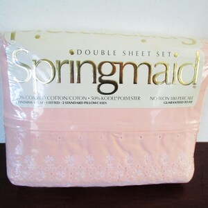 Op de afbeelding: Een verpakte dubbele lakenset van Springmaid. De roze lakens hebben een witte bloemen geborduurde rand. De verpakking bevat de tekst "DOUBLE SHEET SET" en "Springmaid". De set bevat een plat laken, een hoeslaken en twee standaard kussenslopen.
