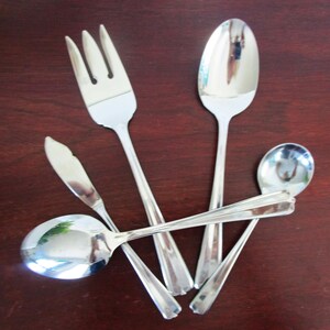 Könnte beinhalten: Eine Sammlung glänzender, silberfarbener Besteckteile, darunter eine Gabel, Löffel und ein Buttermesser. Das Besteck ist auf einer dunkelbraunen Holzoberfläche angeordnet, die das Licht reflektiert. Das Set enthält einen großen und einen kleinen Löffel.