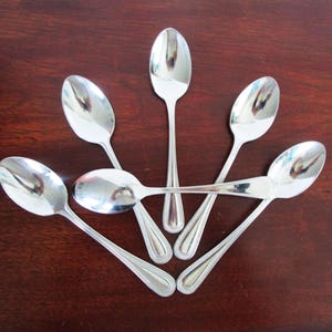 May include: Seven shiny, silver spoons arranged on a dark brown wooden surface. The spoons have a classic design with oval bowls and long, slender handles. The spoons are arranged in a circular pattern.