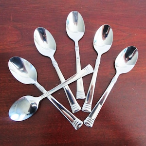May include: Six shiny, stainless steel spoons arranged on a dark wood surface. Each spoon has a rounded bowl and a long handle with a decorative, ridged design near the base. The spoons reflect the surrounding environment.