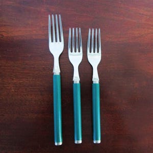 May include: A set of three stainless steel forks with solid teal-colored cylindrical handles. The set includes one larger fork on the left and two smaller forks of identical size positioned to the right, all featuring four tines.