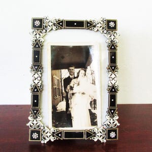 Puede incluir: Un marco de fotos rectangular ornamentado con una fotografía vintage. El marco es dorado con detalles de esmalte negro y pedrería transparente. La fotografía es de una pareja, probablemente una foto de boda, en blanco y negro.