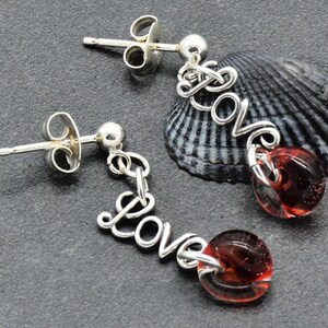 May include: Silver-toned dangle earrings. Each earring features the word "Love" in a cursive script, with a red glass bead at the bottom. The earrings are displayed on a dark gray surface with a seashell in the background.