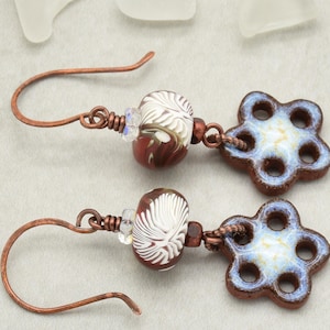 May include: Handmade earrings featuring copper ear wires, brown and white patterned beads, and flower-shaped ceramic charms with blue and white accents. The earrings are displayed on a neutral background, showcasing their intricate details and craftsmanship.