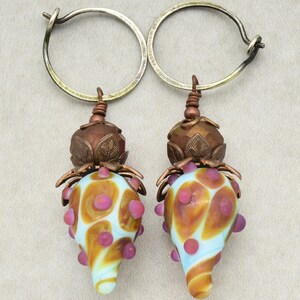 May include: A pair of handmade earrings featuring a light blue and orange glass teardrop-shaped bead with pink accents. The beads are topped with a copper-colored metal floral design and attached to silver-colored hoop earrings. The earrings have a unique, artistic design.