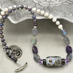 May include: A handcrafted necklace featuring white and dark gray pearls, labradorite, amethyst, and a decorative glass bead with a floral design. The necklace has a silver toggle clasp and a unique pendant. The jewelry is displayed on a decorative silver dish.