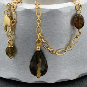 May include: A gold-colored necklace with a chain design. The necklace features a teardrop-shaped, faceted dark brown gemstone pendant and two smaller brown gemstones. A small green gemstone is also present. The necklace is displayed on a textured, silver-colored surface.