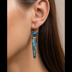 May include: A drop earring featuring a long, tapered, blue and green mottled pendant with a metallic cap. Above the pendant sits a small, round, blue and green marbled bead held by a dark metal wire frame, suspended from a simple metal hook.