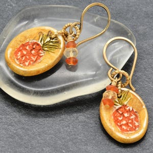 May include: A pair of oval-shaped earrings featuring a hand-painted pineapple design in orange and green on a tan background. Each earring hangs from a gold-toned wire hook and includes small, translucent orange and clear glass beads.