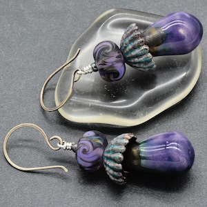 May include: A pair of handmade earrings featuring a teardrop-shaped purple bead with a swirl design, connected to a smaller, round bead with a similar pattern. The earrings have silver-colored ear wires and decorative metal accents.