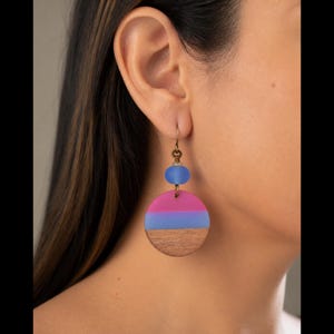 May include: A dangle earring featuring a small, frosted blue bead suspended above a larger, circular pendant. The pendant is divided into three horizontal sections: a top band of bright pink, a middle band of light blue, and a bottom band of wood.