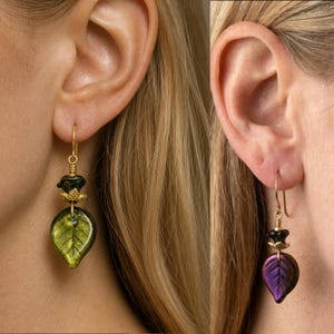 May include: A pair of drop earrings featuring gold-tone wire hooks and small gold-tone caps. One earring displays a translucent green leaf-shaped glass bead, while the other earring features a shimmering purple leaf-shaped glass bead.