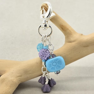 May include: A silver-tone keychain with a lobster clasp, adorned with a cluster of beads. The beads include a light blue flower-shaped bead, a purple crystal-covered bead, and several purple flower-shaped beads. The keychain is hanging on a piece of driftwood.