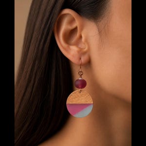 May include: A drop earring featuring a copper-toned wire hook, a small translucent deep red bead, and a circular pendant. The pendant is divided into a textured metallic copper top half and a smooth bottom half with diagonal pink and gray stripes.