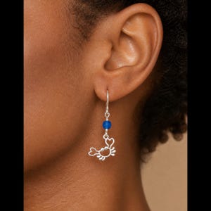May include: A silver dangle earring featuring a small, stylized crab charm suspended from a thin wire hook. Above the crab charm, a single, round, vibrant blue bead is integrated into the silver wire, adding a pop of color to the design.