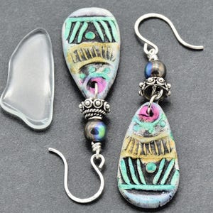May include: Artisan earrings with a unique design. The earrings feature teardrop-shaped, hand-painted beads in shades of blue, yellow, and pink, accented with silver-toned metal beads and ear wires. The design includes geometric patterns and a clear, teardrop-shaped accent piece.