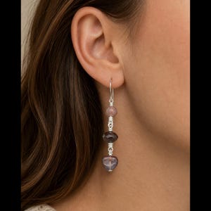 May include: A drop earring featuring three faceted, translucent purple and mauve beads arranged vertically. The beads are connected by silver-toned wire wrapping, with the bottom bead shaped like a heart. The earring hangs from a silver-toned hook.