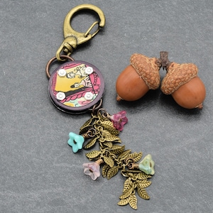 May include: A decorative keychain with a round, patterned charm, a bronze-colored floral and leaf charm, and a bronze-colored clasp. Two acorns are next to the keychain. The keychain has a yellow, pink, and white design.