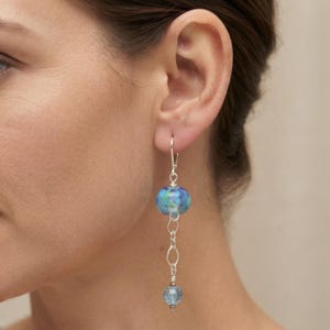 May include: A drop earring featuring a silver-toned leverback hook, a larger round glass bead with swirling blue and teal patterns, a silver-toned metal link chain, and a smaller, translucent light blue faceted glass bead at the bottom.