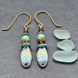 May include: Dangle earrings with gold-colored hooks. Each earring features a feather-shaped ceramic bead in blue, tan, and purple, accented with turquoise beads, gold-colored leaf charms, and dark red beads. Three pieces of sea glass are also visible.