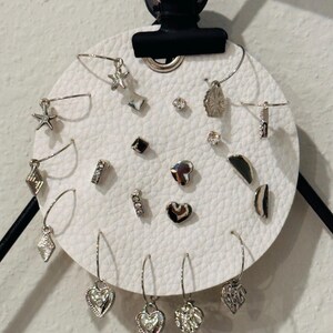 May include: A collection of silver-toned earrings displayed on a white, circular, textured surface. The earrings feature various designs, including stars, hearts, and geometric shapes. Some are studs, while others are dangling hoops.