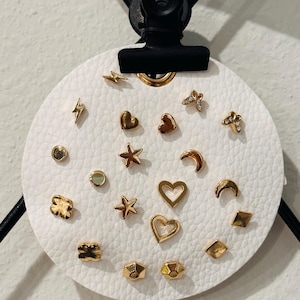 May include: A collection of gold-colored stud earrings in various shapes, including hearts, stars, lightning bolts, and geometric designs. The earrings are displayed on a white, circular, textured surface, held by a black clip.