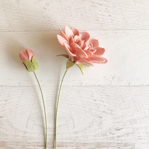 May include: Two felt flowers, one pink and one a closed bud, on a white wooden background.