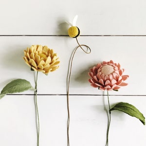 Op de afbeelding: Drie vilten bloemen op stelen tegen een witte achtergrond. Eén is geel, één is roze en de derde is een grillige bij. De bloemen hebben groene bladeren. De achtergrond is een wit houten oppervlak.