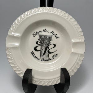 May include: White ceramic ashtray with a black and white image of the Eden Roc Hotel in Miami Beach, Florida. The image includes the hotel's logo, a column with a capital, and the text "Eden Roc Hotel Miami Beach, Fla."
