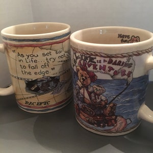 May include: Two ceramic mugs with a nautical theme. One mug features a map of the Pacific Ocean with the quote "As you set sail in life... try not to fall off the edge." The other mug features a cartoon bear wearing a sailor hat and holding a fishing rod with the quote "Life is a daring adventure".