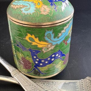 May include: A cylindrical green cloisonné jar with a matching lid, featuring colorful enamel depictions of a blue deer, a yellow dragon, and floral motifs outlined in gold wire. Two silver-toned metal utensils with intricate engraved patterns lie below.