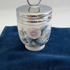 May include: A white ceramic egg cup with a floral design in shades of purple, blue, and yellow. The egg cup has a silver-colored metal lid with a ring handle. The cup is sitting on a dark blue surface.