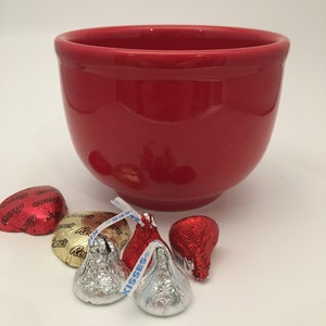 May include: A red ceramic bowl with a smooth, glossy finish.  There are five foil-wrapped chocolate kisses in the foreground, two red, two silver, and one gold.