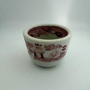 May include: A small, white ceramic cup with a red, detailed Willow pattern design. The cup has a rounded shape and a thick rim. The pattern features a landscape with buildings, trees, and figures. The cup is likely vintage.