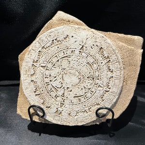 May include: A white stone replica of the Aztec calendar, also known as the Sun Stone, with intricate carvings and glyphs. The stone is on a black metal stand.