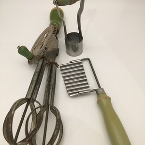 May include: Three vintage kitchen tools with green wooden handles. A hand-crank egg beater, a metal cookie cutter, and a wavy-edged pastry cutter.