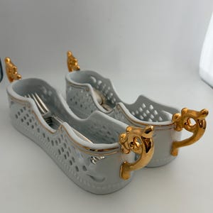 May include: Two white porcelain utensil holders with gold accents. Each holder has a pierced design and a gold handle. The holders are designed to hold forks and spoons. The holders are on a white surface.