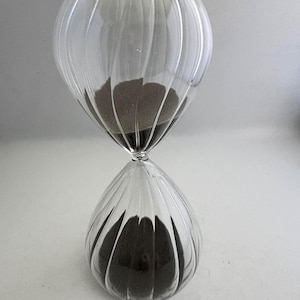 May include: A clear glass hourglass featuring a ribbed, vertical texture on the exterior. The hourglass contains dark brown sand, with a portion currently resting in the top bulb and the majority of the sand collected in the bottom bulb.