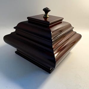May include: A dark brown, wooden box with a square, tiered design and a decorative brass knob on the lid. The box has a glossy finish and is likely used for storage or display.