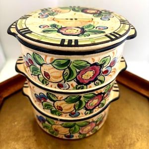 May include: Stacked ceramic serving bowls with lids. The bowls are cream-colored with a floral design in shades of red, blue, green, and yellow. The lids have a black border and a handle. The bowls are stacked on a gold-colored tray.